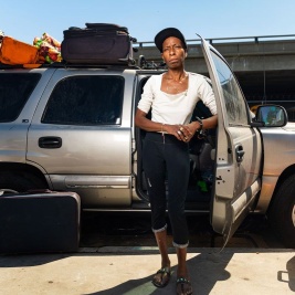 Portrait of a Black unhoused woman standing in front of her gray SUV, elbow propped against a door
