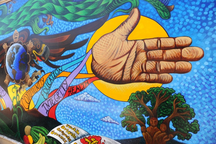 A section of a vivid, painted mural showing an outstretched open hand in front of the sun