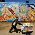 A vendor pushes a paletas cart in front of a colorful mural near Fruitvale, California