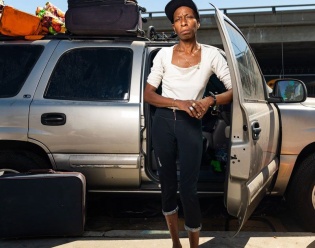 Portrait of a Black unhoused woman standing in front of her gray SUV, elbow propped against a door